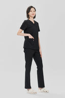 Model wearing Black Avison Scrub Top by Good Uniform with a hand in the pocket, full front view slightly from the side