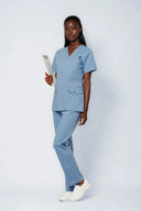 Model wearing Soft Blue Avison Scrub Top by Good Uniform in a clinical setting, full front view slightly form the side