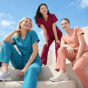 Three women in matching medical scrubs sit and stand on outdoor concrete steps under a blue sky. One wears teal scrubs, one wears burgundy scrubs, and one wears peach-pink scrubs.