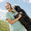 A smiling woman in light green medical scrubs holds and kisses a fluffy black-and-white dog outdoors against a blue sky background.